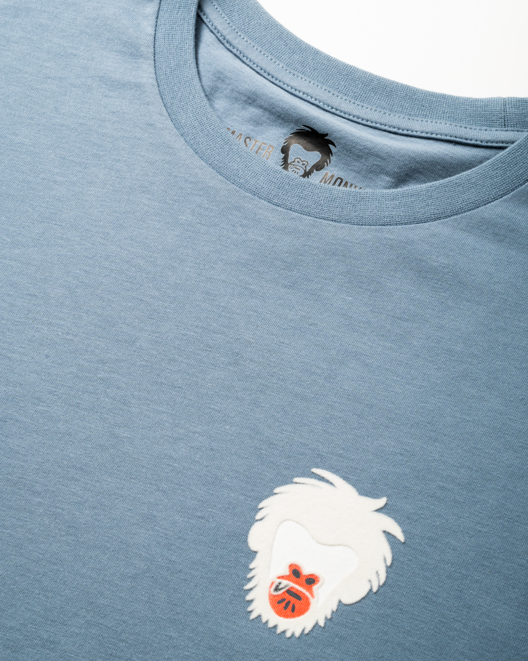 Close-up of a blue t-shirt with a white embroidered Master Monkey logo on a light gray background #color_steel-blue