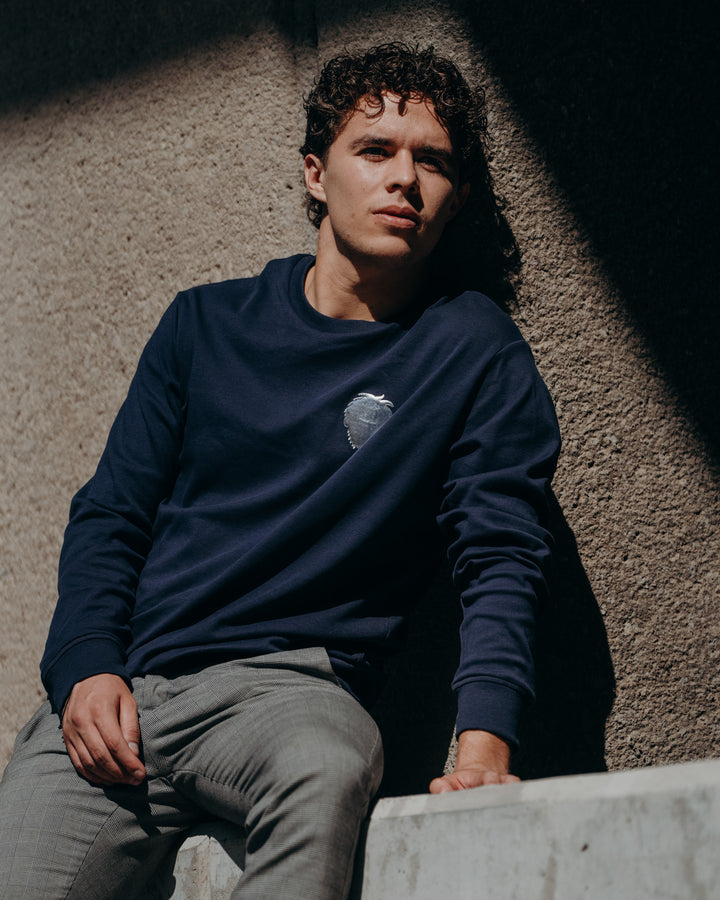Person wearing a navy blue sweatshirt with a logo, sitting against a textured wall. #color_midnight