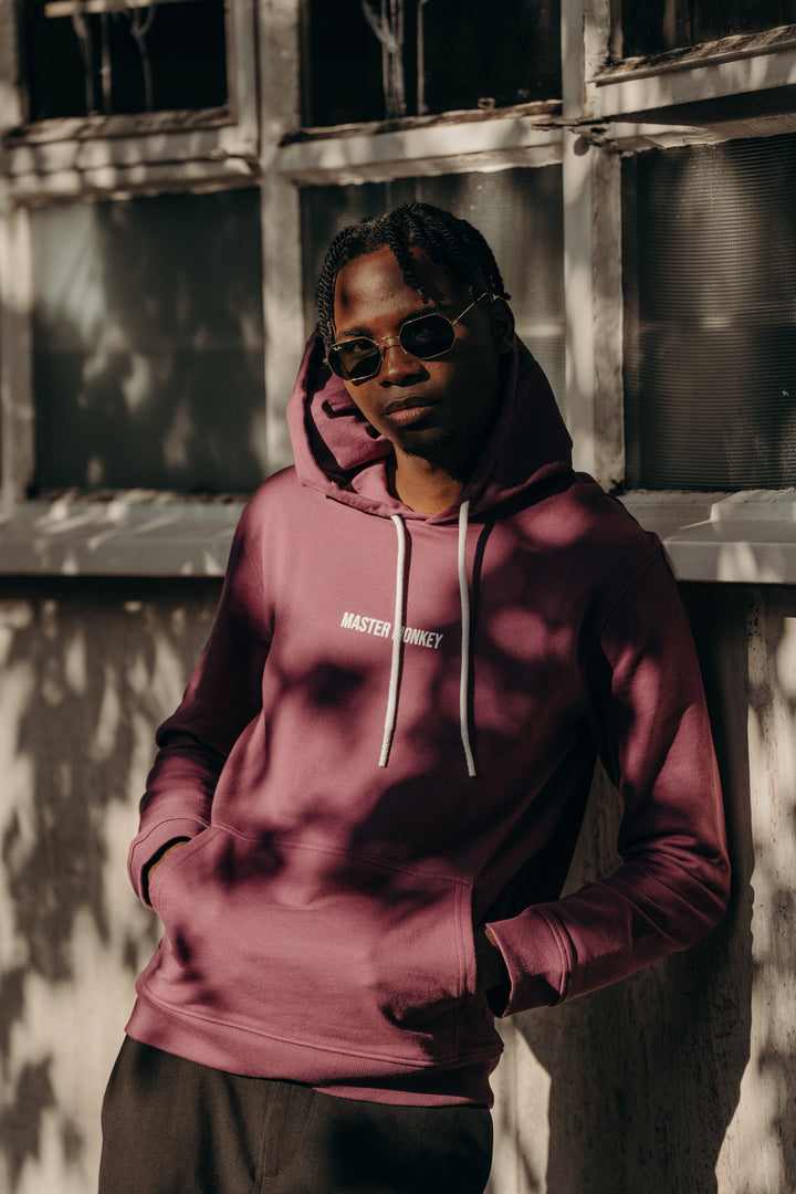 Person wearing a maroon hoodie with 'Master Disguise' text, standing against a textured wall. #color_berry