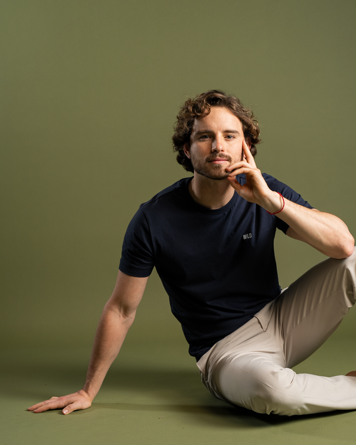 Man sitting on a green background wearing a navy blue t-shirt and beige pants. #color_midnight